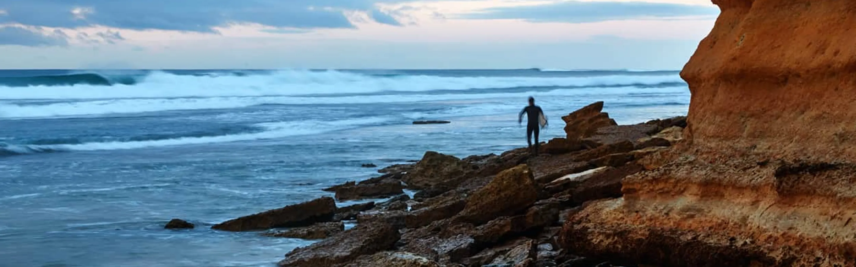 A surf boarder walks out on the rocks at the ocean