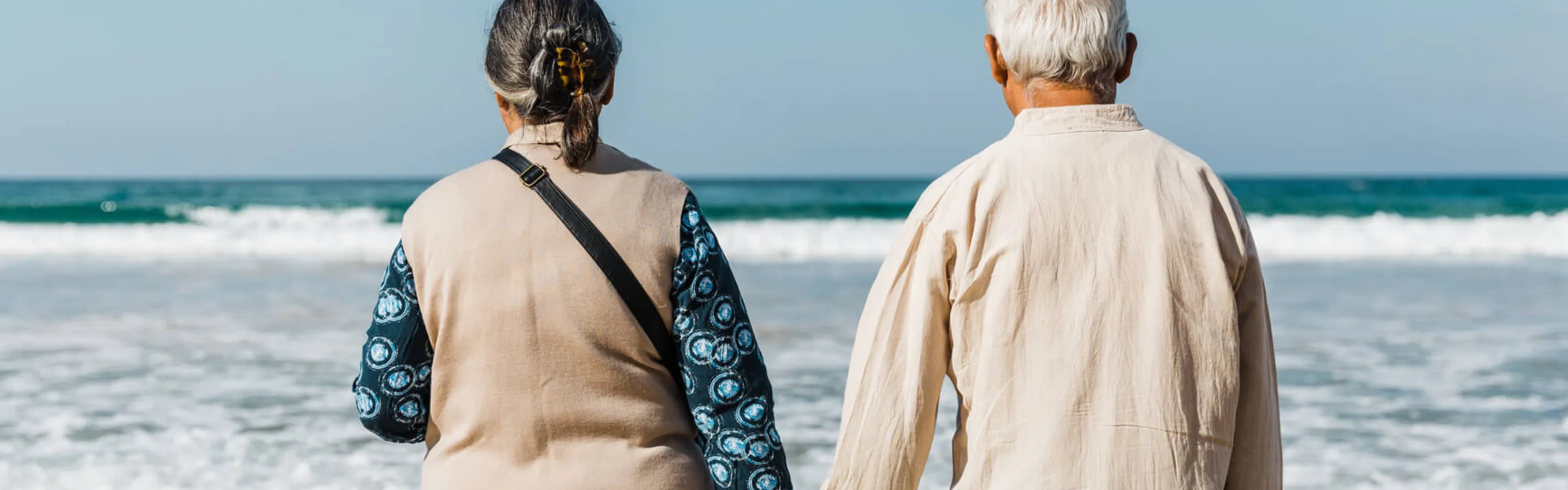 An older couple holding hands, their back is to the camera as they look out at the ocean from a white sand beach