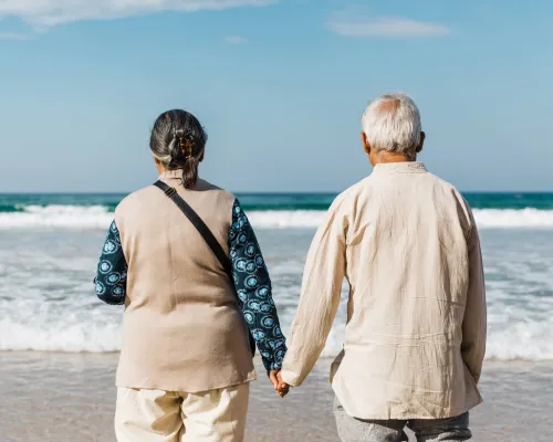 An older couple holding hands, their back is to the camera as they look out at the ocean from a white sand beach